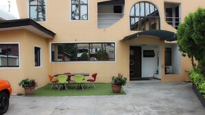 Ein Gebäude mit flachem Dach, großen Fenstern und einem überdachten Eingang. Es gibt eine kleine Außenbereich-Sitzgelegenheit mit Stühlen und Tischen.