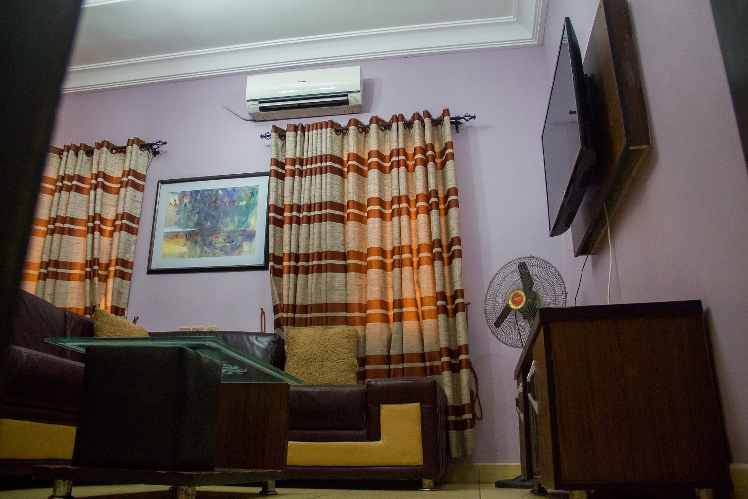 A living room with a sofa, a glass coffee table, a wooden cabinet, a wall-mounted TV, and a standing fan.