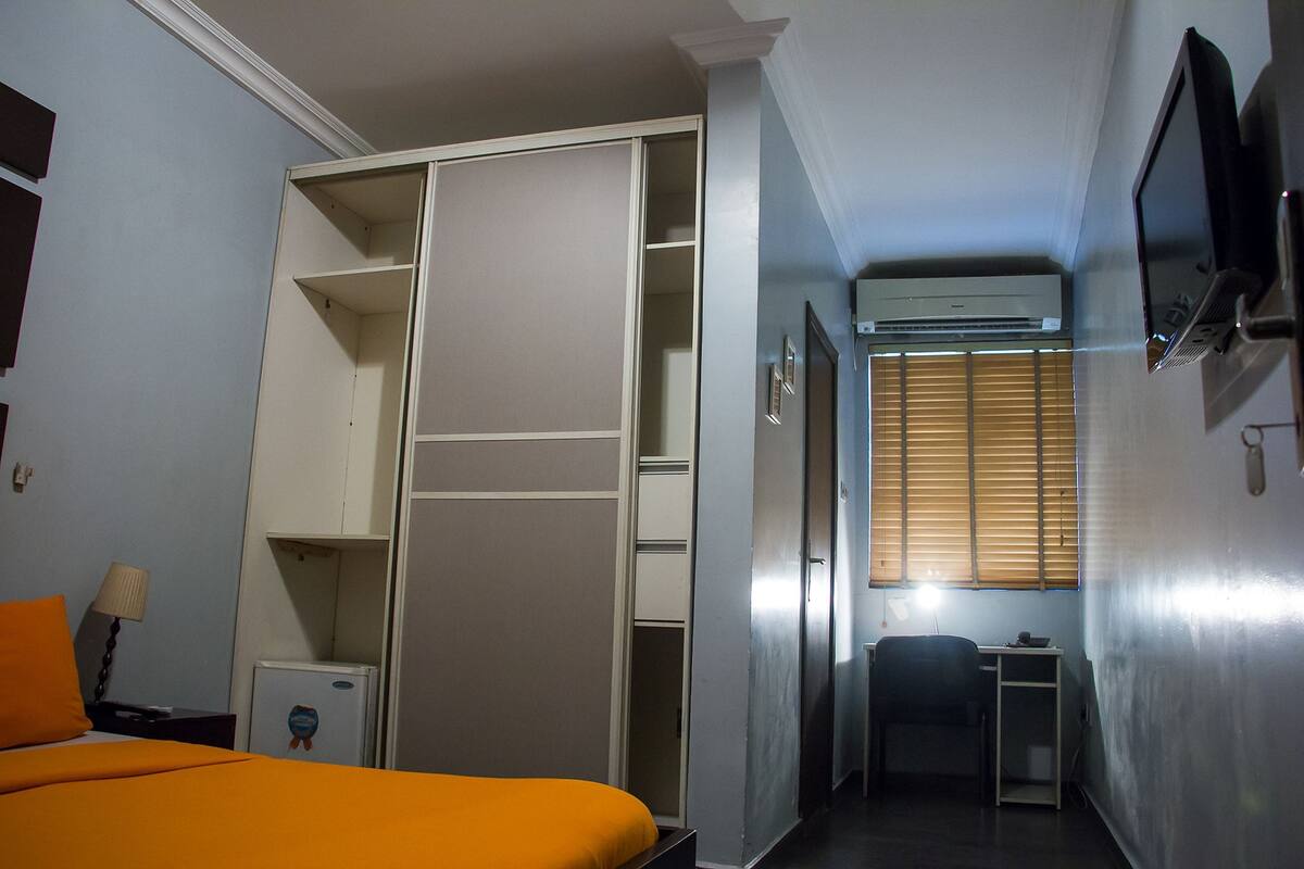 A hotel room with a bed, a desk, a wardrobe, and an air conditioning unit.