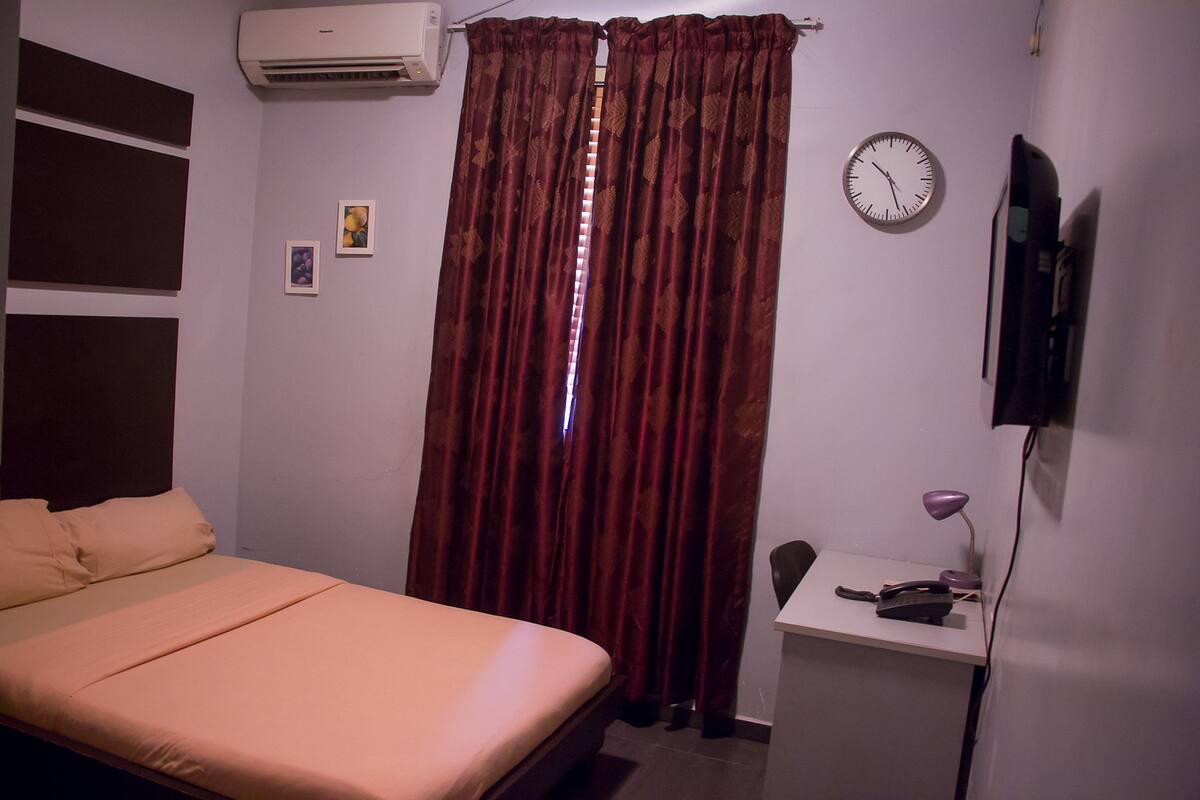 A bedroom with a bed, a desk, a chair, a clock, and a television on the wall.