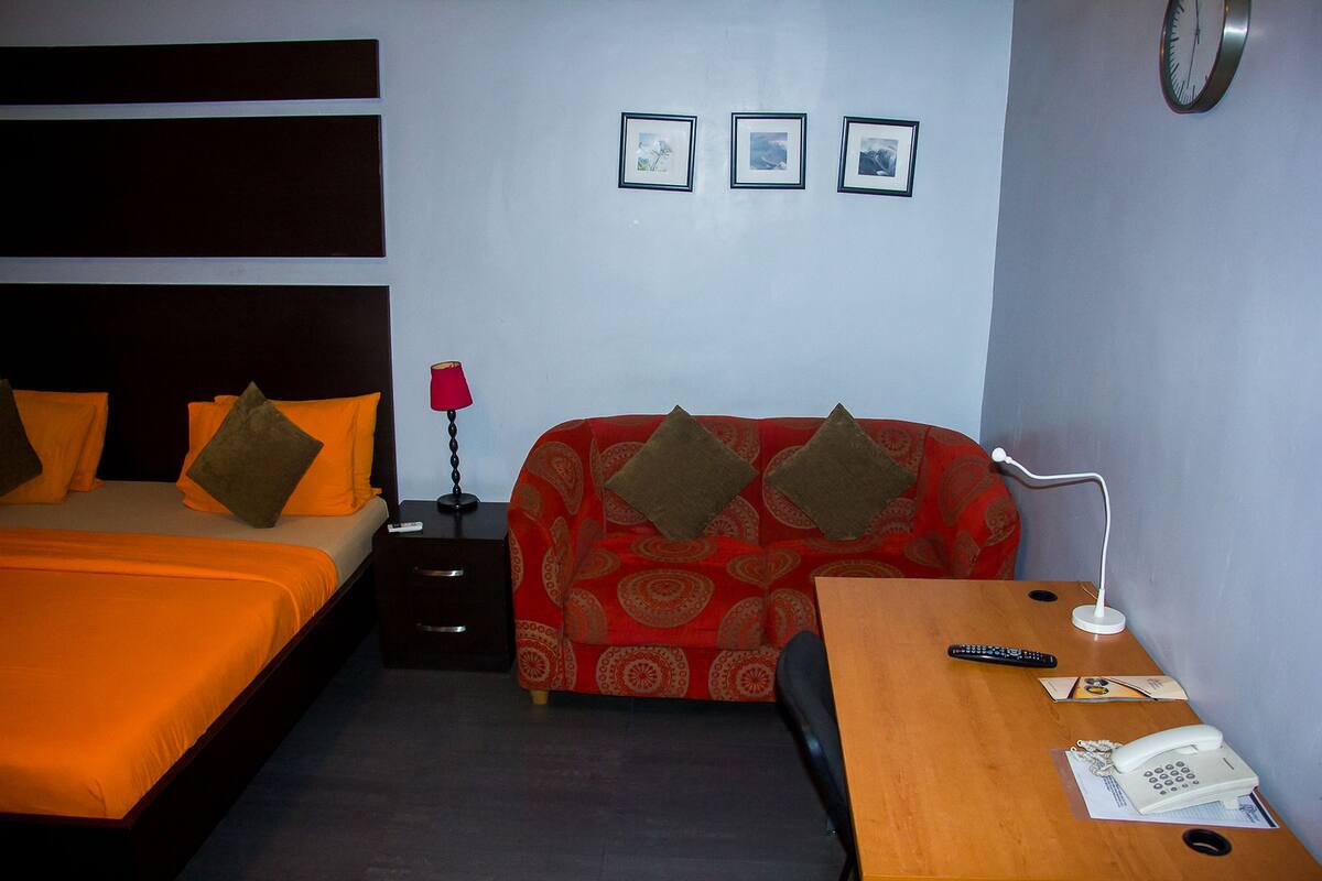 A hotel room with a bed, a sofa, a desk with a lamp, a phone, and a clock on the wall.
