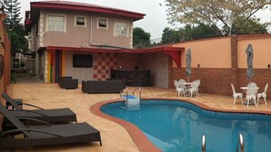 A swimming pool with lounge chairs and tables, surrounded by a brick wall and a building with a red roof. - Porschester Hotel and Suite (Lagos)