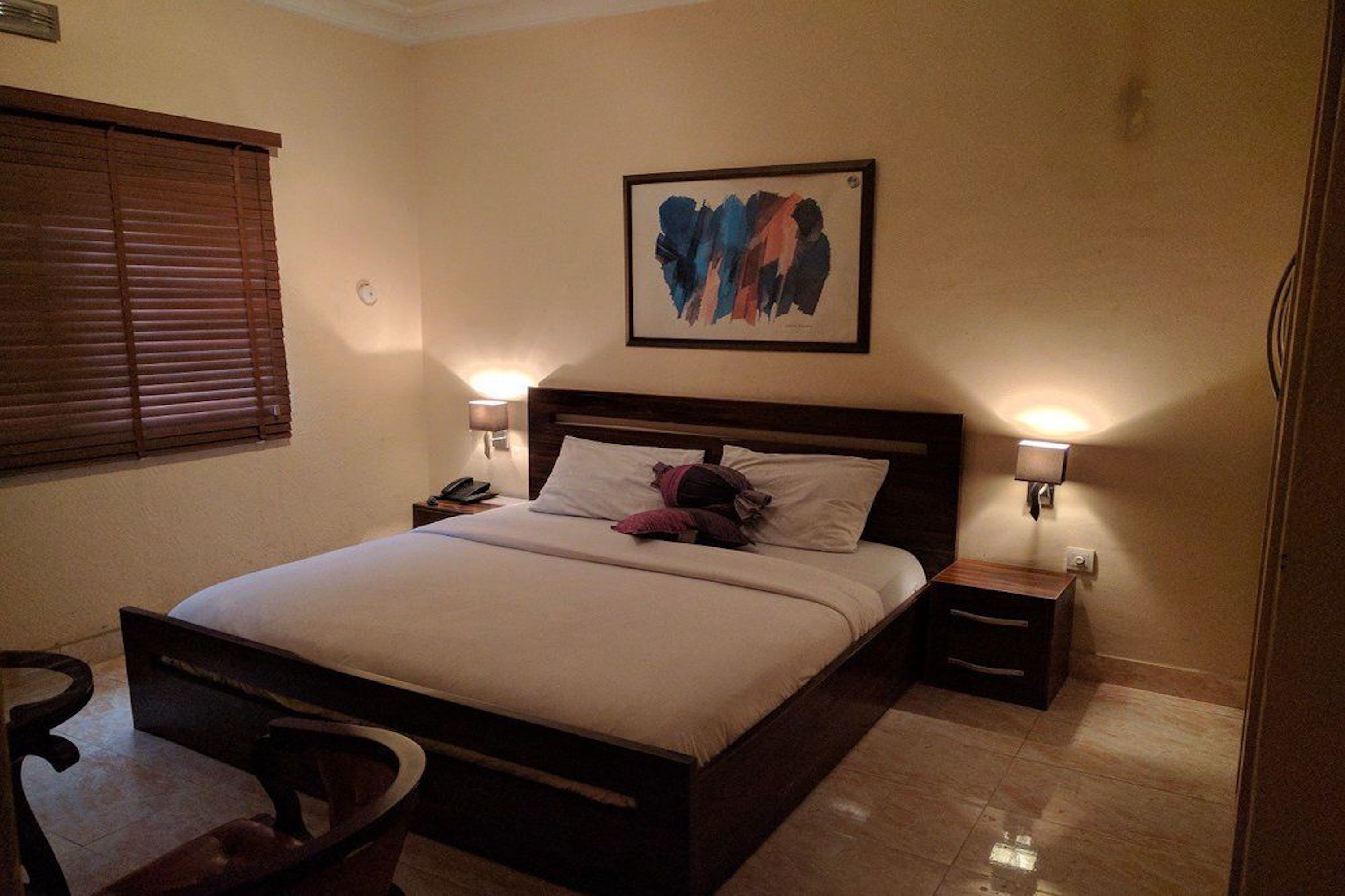 A hotel room with a bed, bedside lamps, a nightstand, a chair, and a painting on the wall.