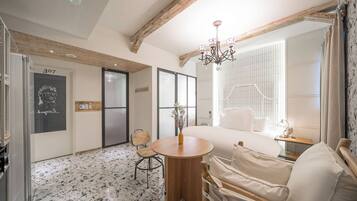 Une chambre d’hôtel avec un lit, une table ronde en bois, une chaise, une petite table avec un vase, un miroir et une porte avec un numéro.