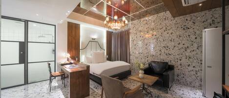 A hotel room with a bed, a desk, a chair, a small table with a vase, a black leather sofa, and a refrigerator.