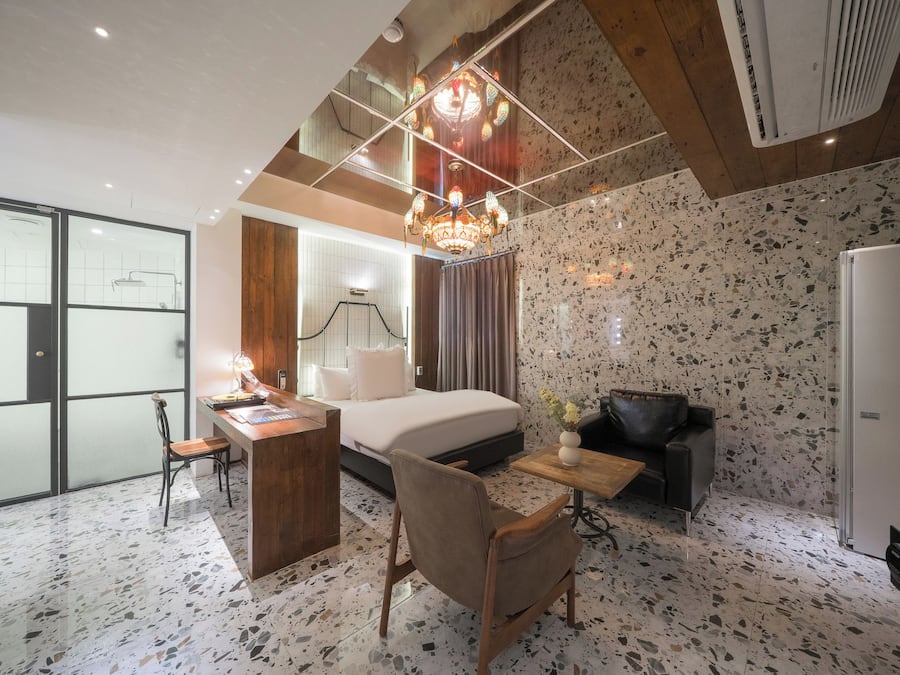A hotel room with a bed, a desk, a chair, a small table with a vase, a black leather sofa, and a refrigerator.