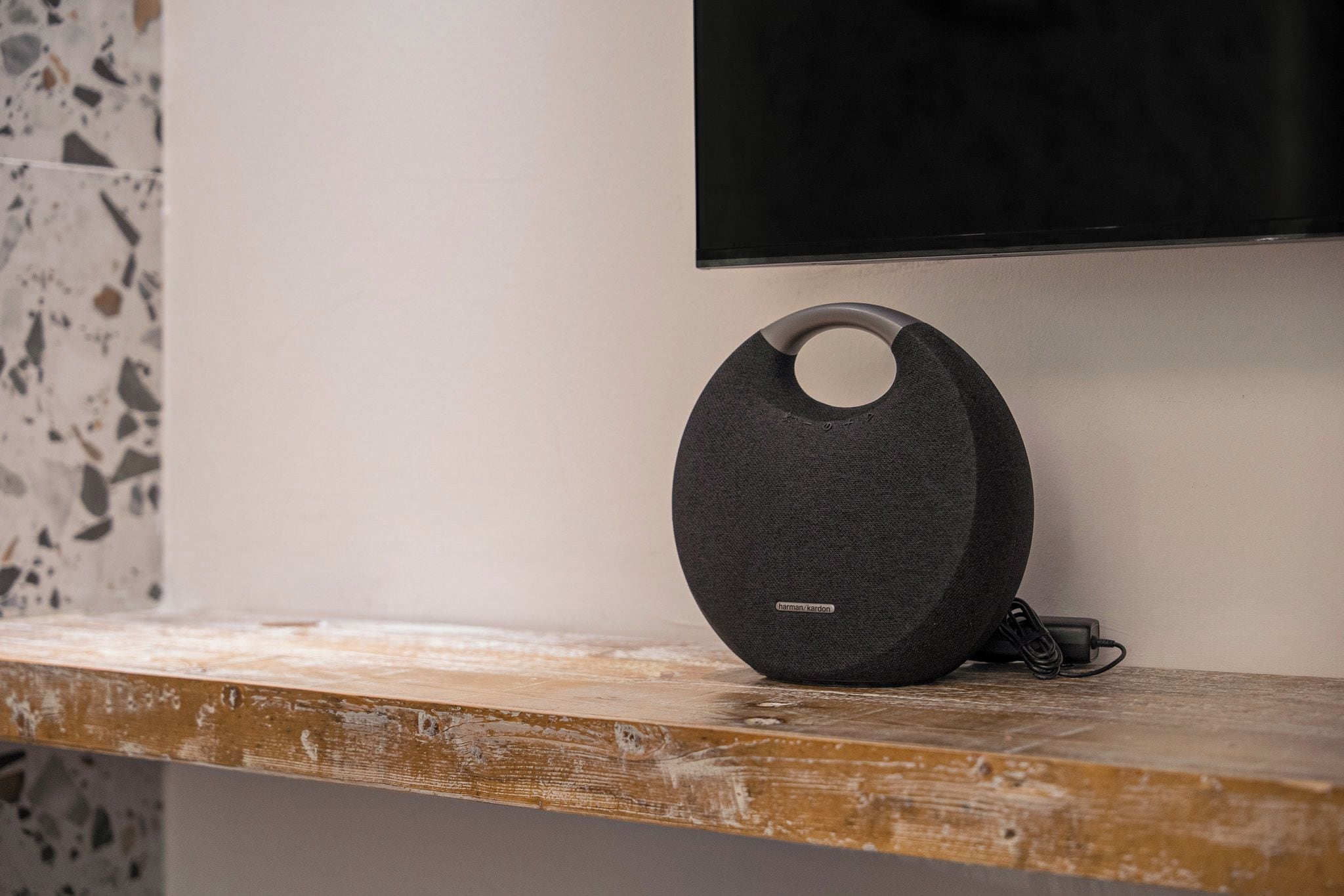 A black speaker with a circular design on a wooden surface, a television mounted on the wall, and a terrazzo floor.