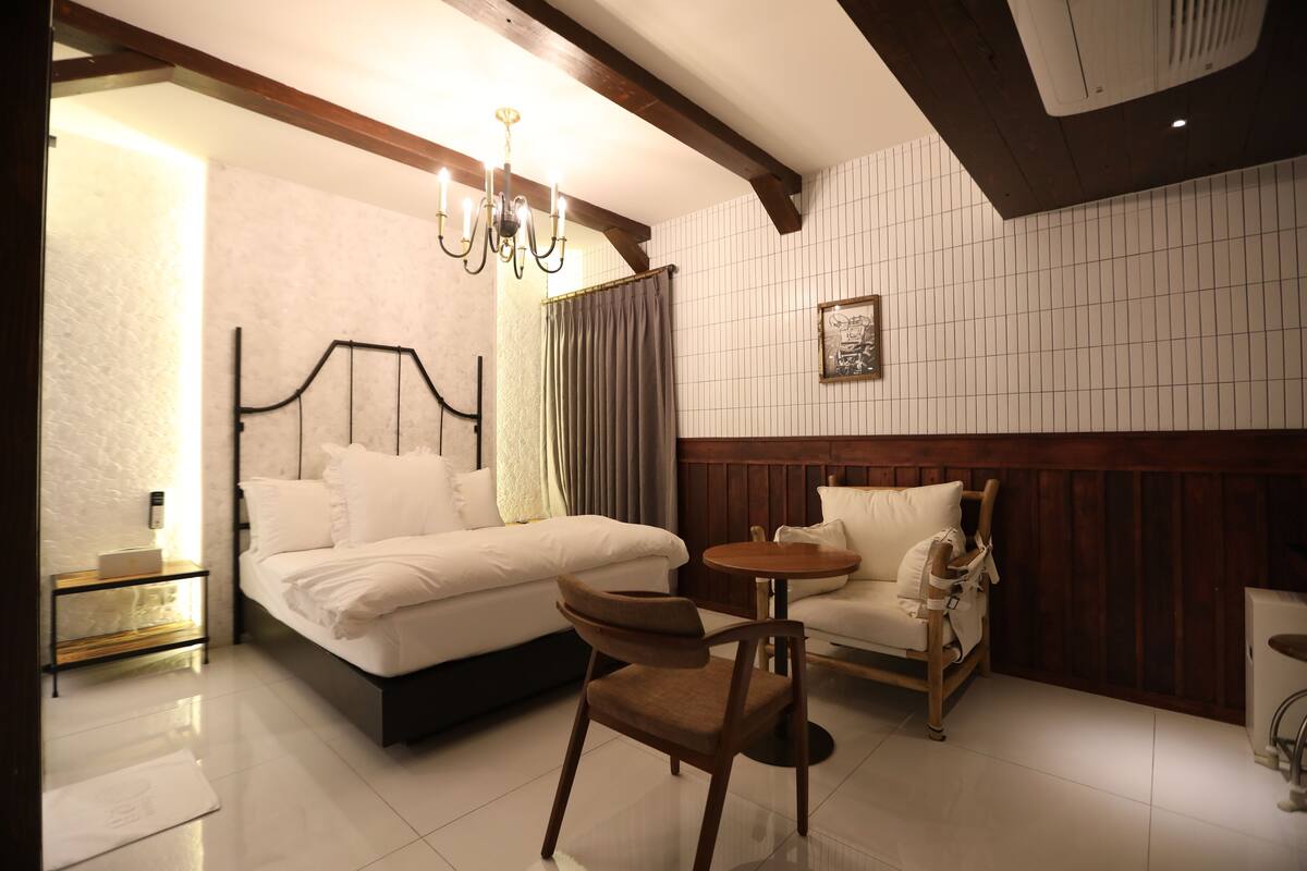 A hotel room with a bed, a chair, a small table, a nightstand, a chandelier, and a picture on the wall.