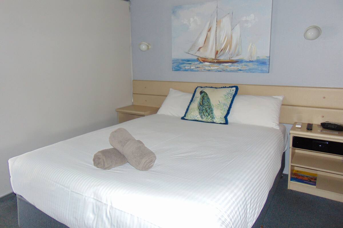 A neatly made bed with two towels, a painting of a sailboat on the wall, and a bedside table with a phone and remote control.