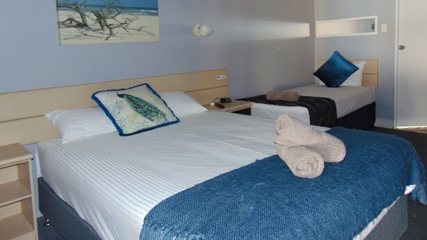 A hotel room with two beds, a painting of a beach, a nightstand, and a door. - Rockhampton Court Motor Inn (Rockhampton)