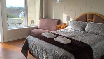 A bedroom with a large window, a bed with a patterned bedspread, a chair, and a nightstand with a lamp.