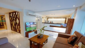 A modern living room with a sofa, a coffee table with a plant, a TV, and a kitchen area in the background. - Emerald Boutique Hotel (Legazpi)