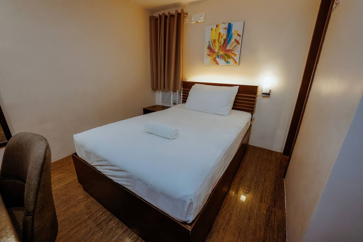 A neatly made bed with a white comforter and a rolled towel, a wooden headboard, a bedside table, a chair, and a colorful abstract painting on the wall.