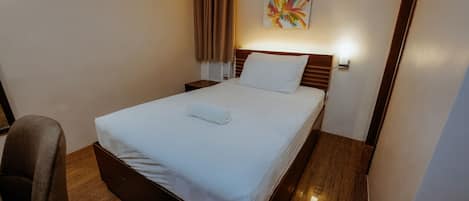 A neatly made bed with a white comforter and a rolled towel, a wooden headboard, a bedside table, a chair, and a colorful abstract painting on the wall.