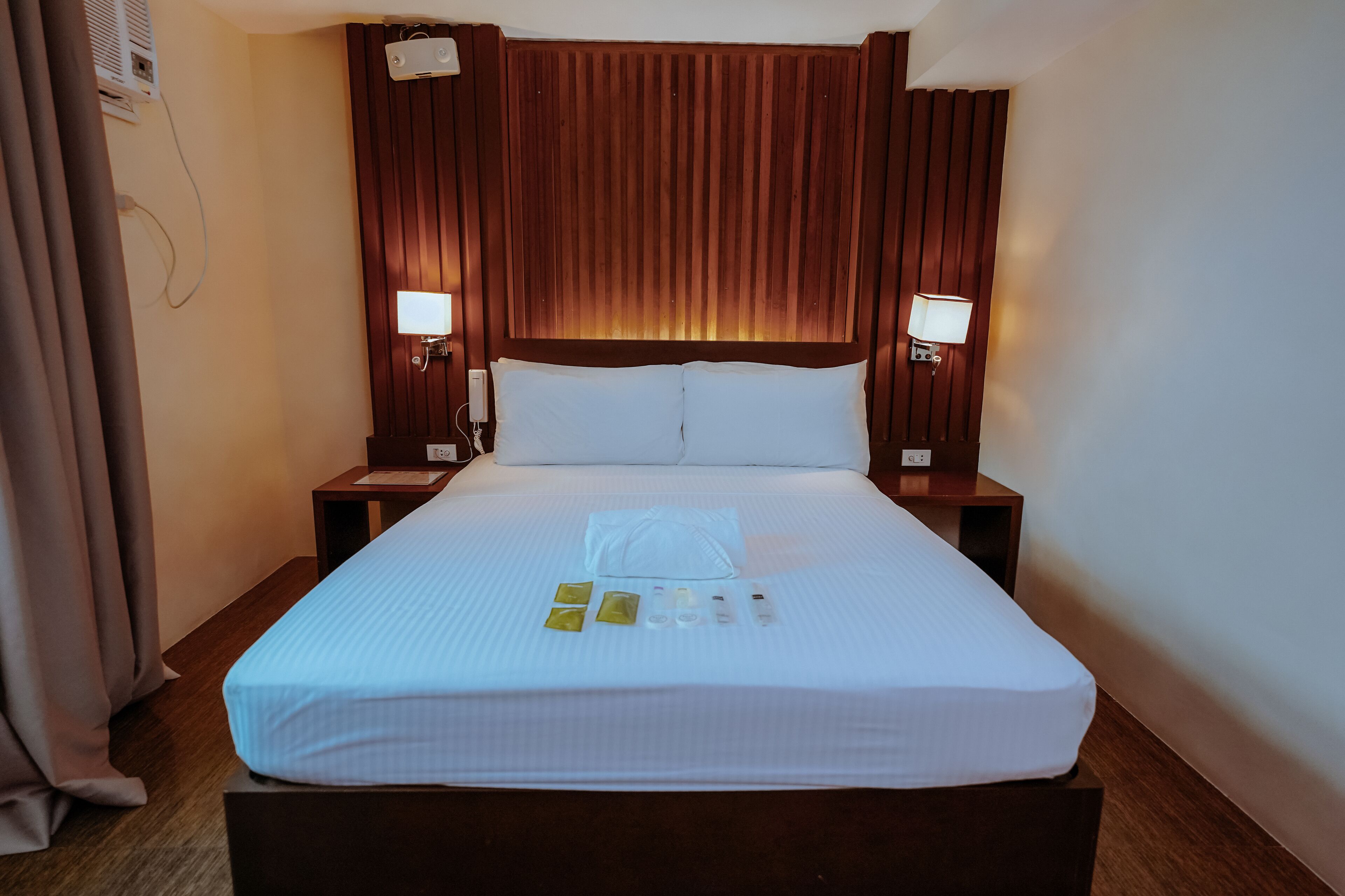 Une chambre d’hôtel avec un grand lit, deux tables de chevet avec des lampes, une tête de lit en bois et une fenêtre avec des rideaux.