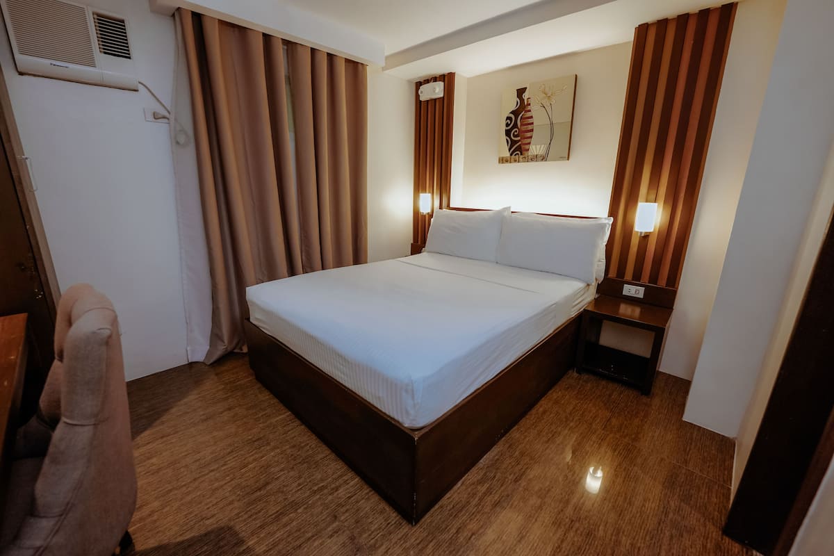 A hotel room with a large bed, wooden floor, curtains, a nightstand with a lamp, and a wall-mounted air conditioning unit.