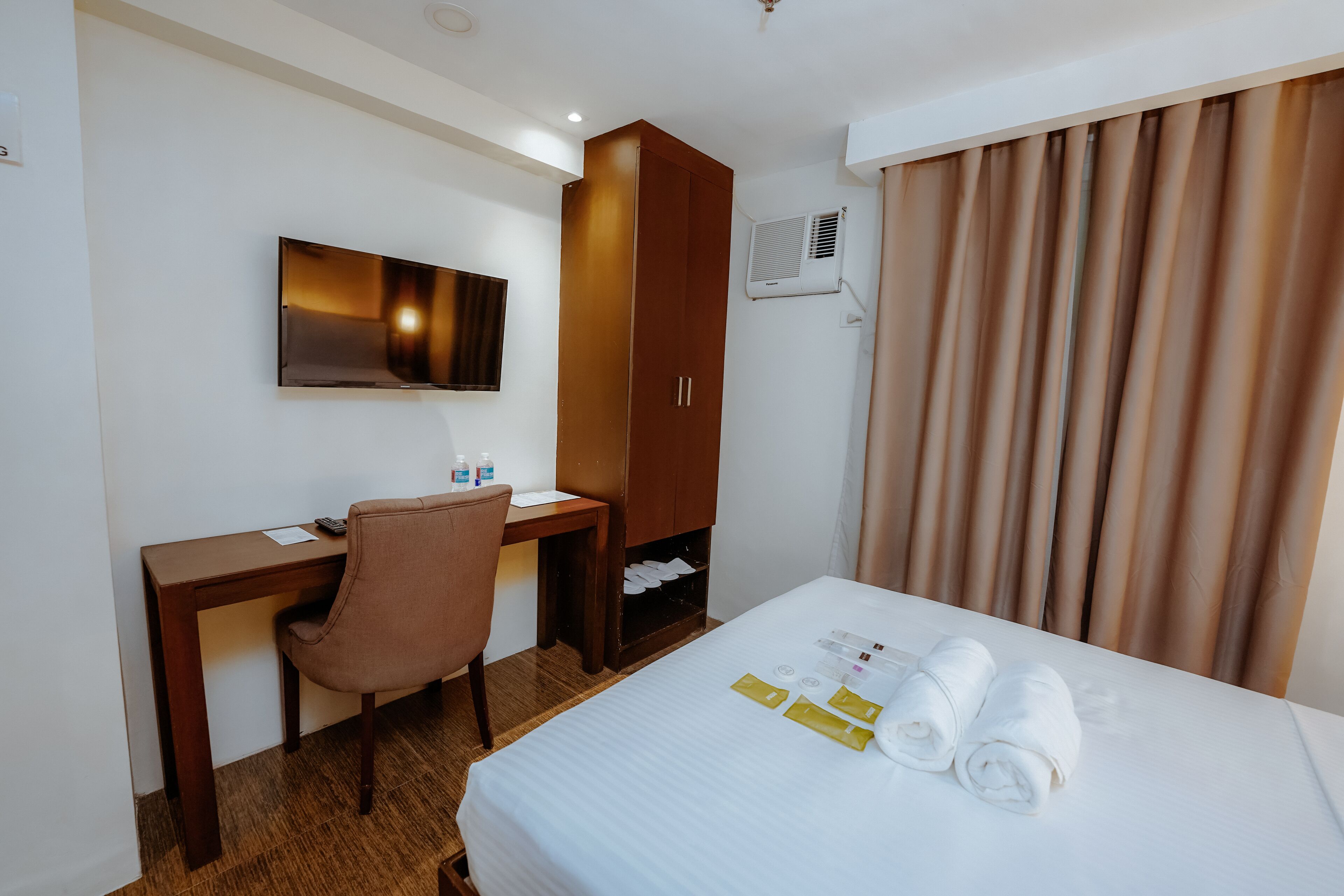 A hotel room with a bed, a desk, a chair, a wardrobe, a flat-screen TV, and a window with curtains.