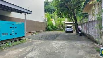 Un callejón estrecho con un coche blanco al final, rodeado de follaje y un cuadro eléctrico azul a la izquierda.