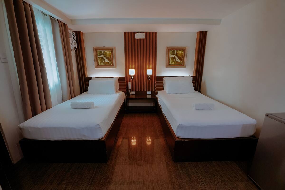 A hotel room with two single beds, a wooden floor, a small table, and two wall-mounted lamps.
