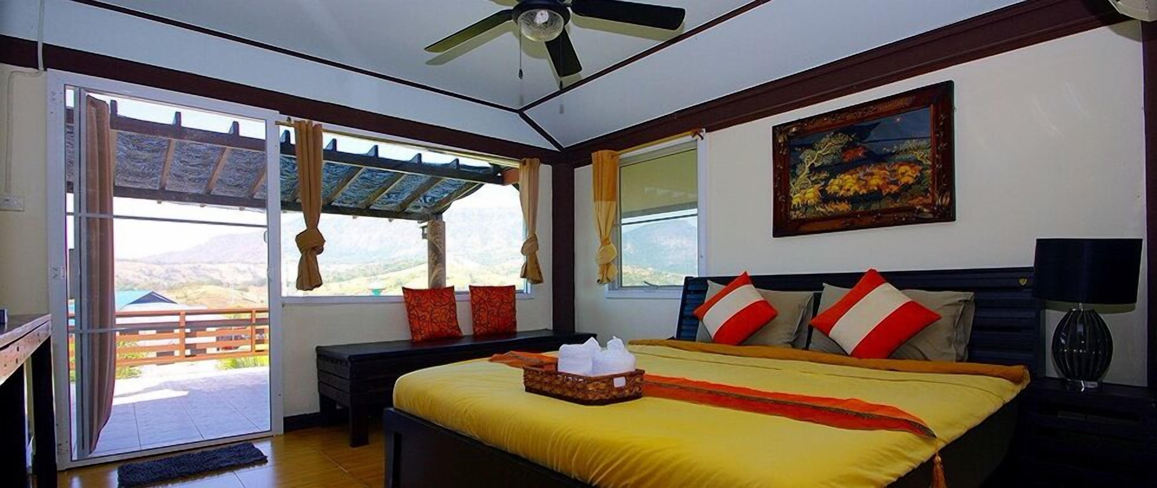 A bedroom with a bed, a ceiling fan, a painting, and a view of the outdoors.