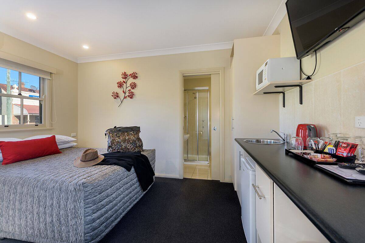A compact hotel room with a bed, a small kitchenette, a shower, and a view of the outside.