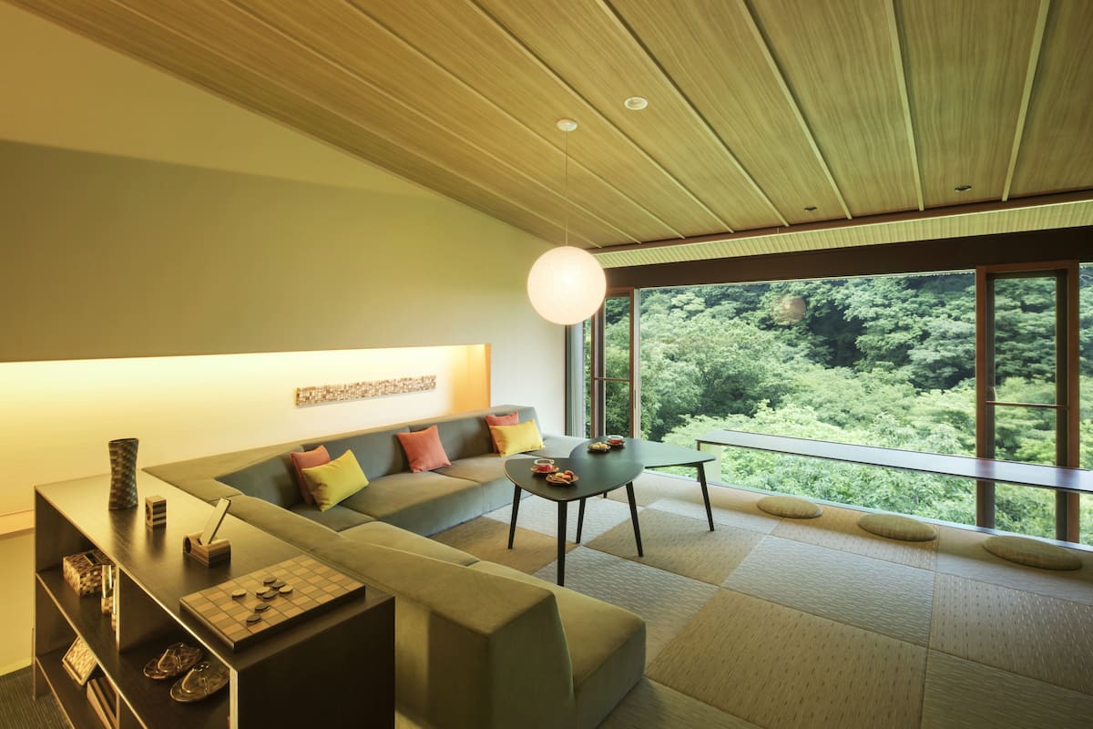 A modern living room with a sectional sofa, a dining table, and a large window offering a view of greenery.