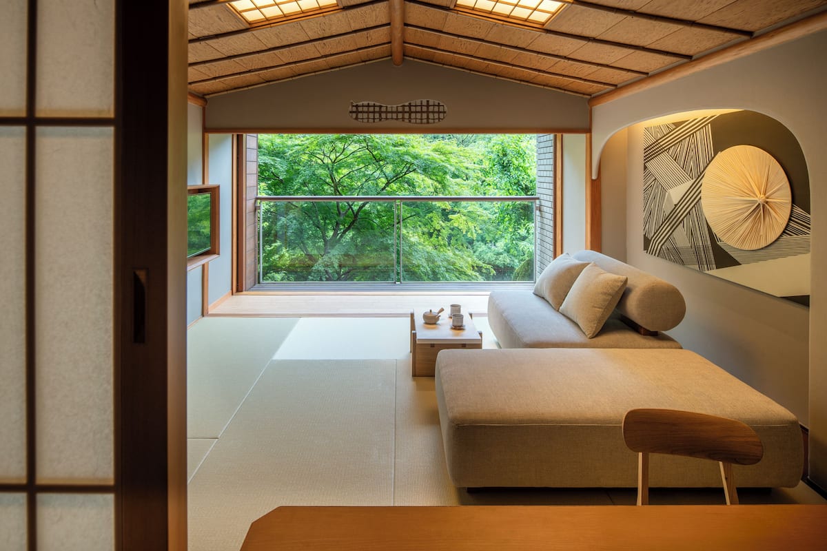 A room with tatami flooring, a sofa, a wooden table, and a view of greenery through large windows.