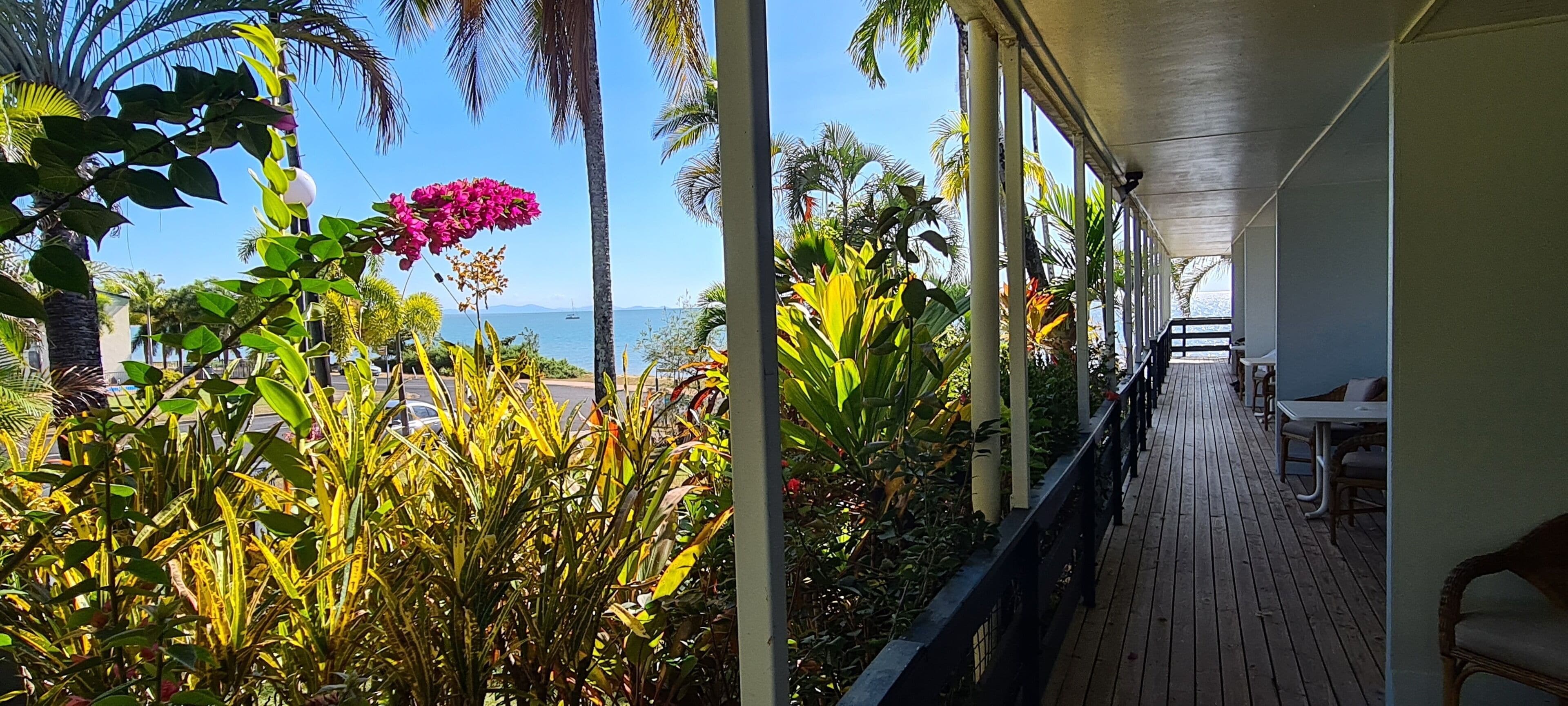 A covered outdoor area with a wooden deck, surrounded by lush tropical plants and palm trees, overlooking a body of water.