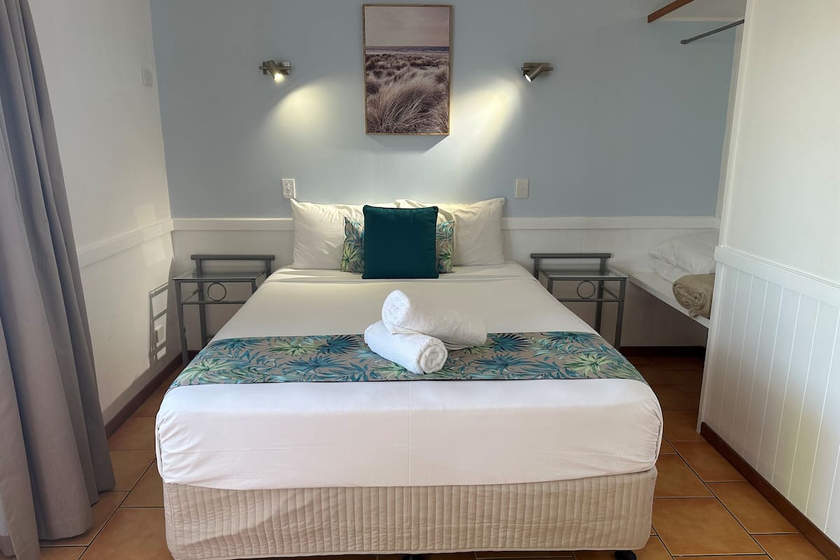 A neatly made bed with a floral bedspread, two bedside tables, a wall-mounted light, and a painting above the bed.