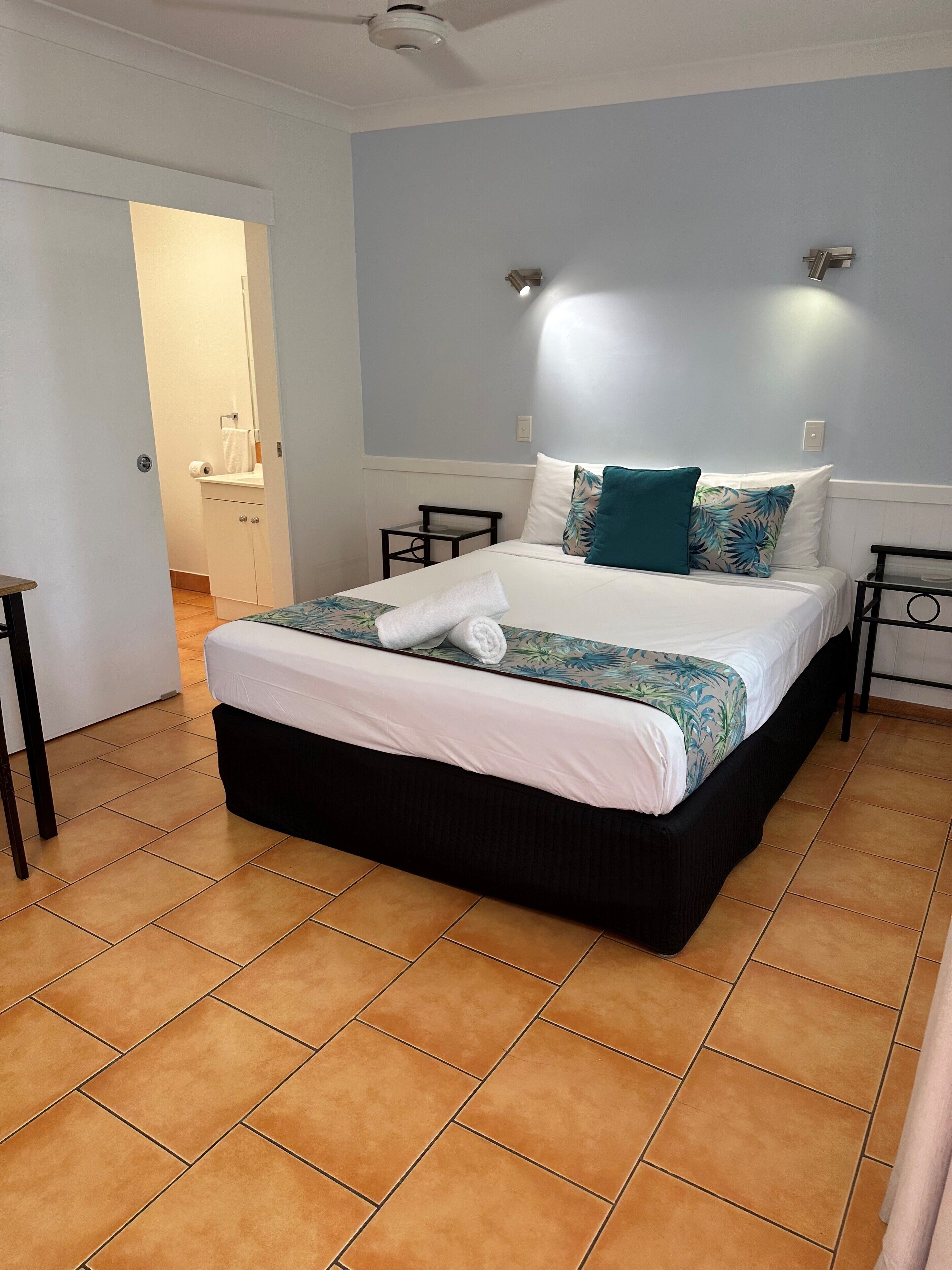 Un dormitorio ordenado con una cama, una mesita de noche, una lámpara en la pared y una puerta que da al baño.