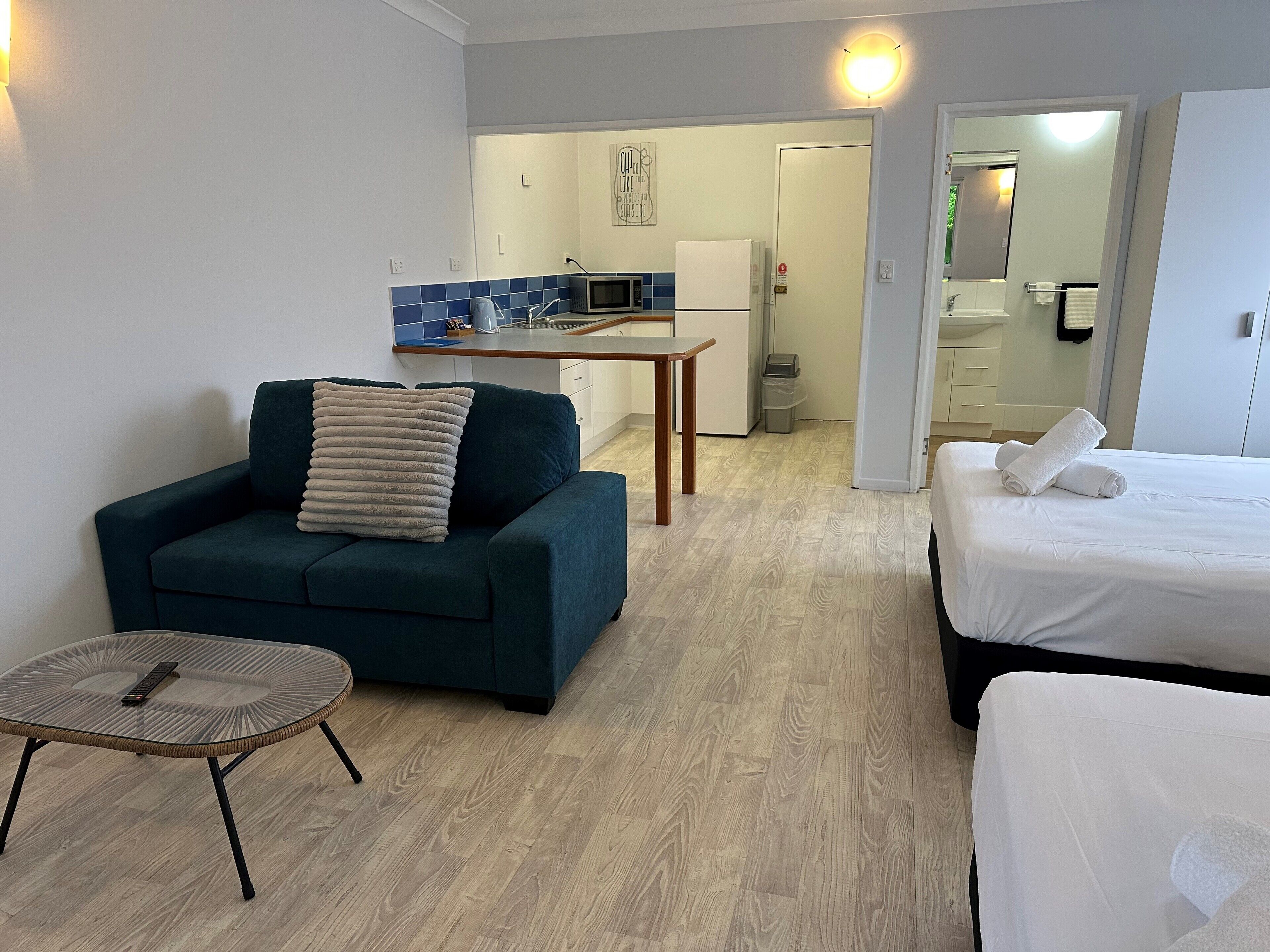 Et kompakt hotelværelse med sofa, lille rundt bord, seng med pænt foldede håndklæder, tekøkken med mikroovn og et badeværelse, der kan ses gennem en åben dør.