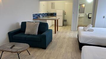 Et kompakt hotelværelse med sofa, lille rundt bord, seng med pænt foldede håndklæder, tekøkken med mikroovn og et badeværelse, der kan ses gennem en åben dør.
