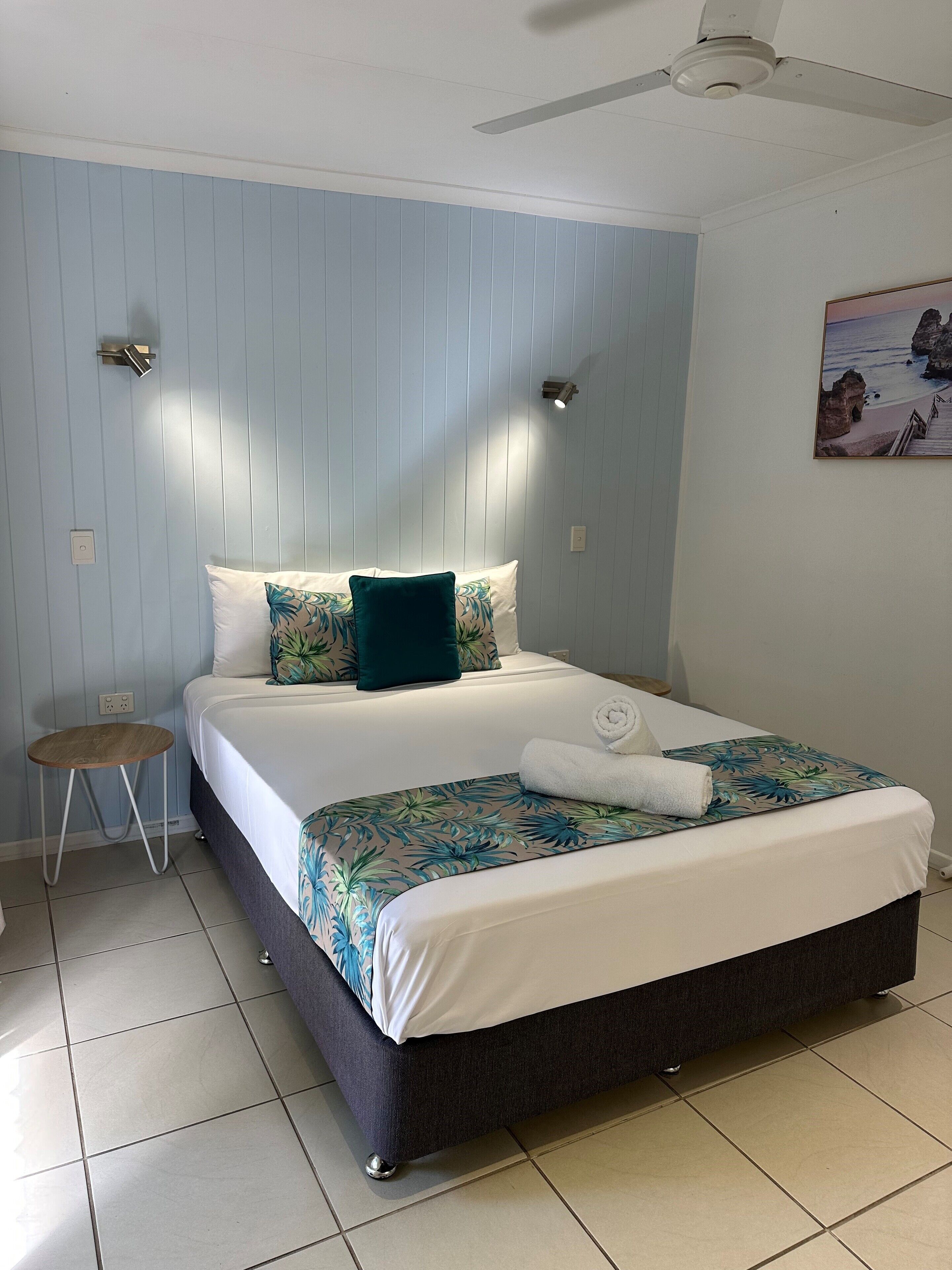 A neatly made bed with a floral bedspread, a small wooden stool, and a ceiling fan.
