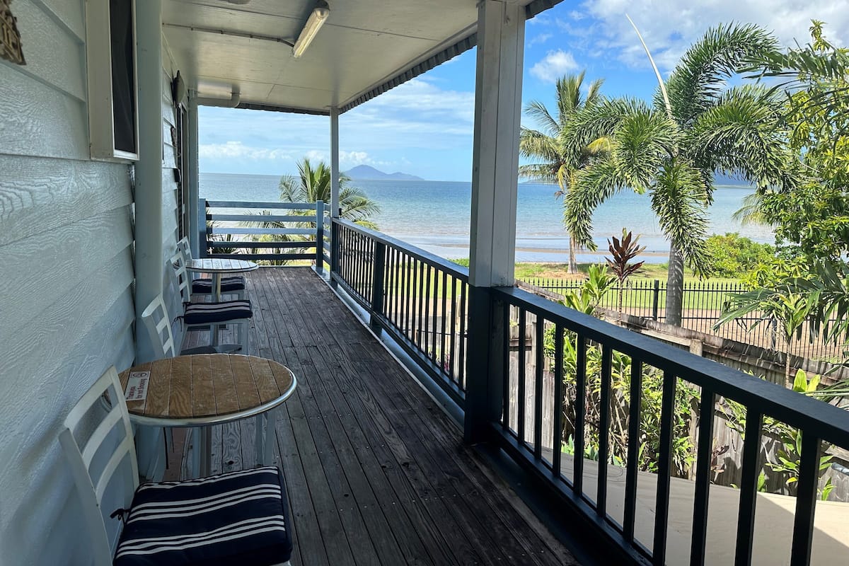 A wooden deck with a table and chairs, overlooking a beach with palm trees and a clear sky.