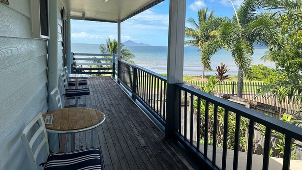 Una terraza de madera con una mesa y sillas, con vista a una playa con palmeras y cielo despejado.