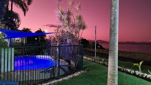Piscina rodeada de una valla, palmeras y vista al océano al atardecer.