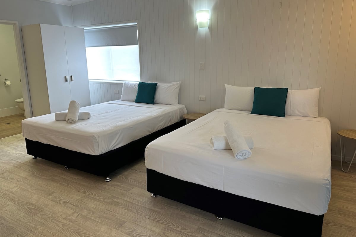 Two single beds with white linens and teal pillows, a small round table, and a window with blinds.