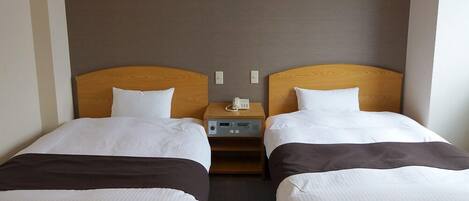 Habitación de hotel con dos camas individuales, una mesita de noche con un teléfono y un pequeño dispositivo electrónico, y una pared lisa.