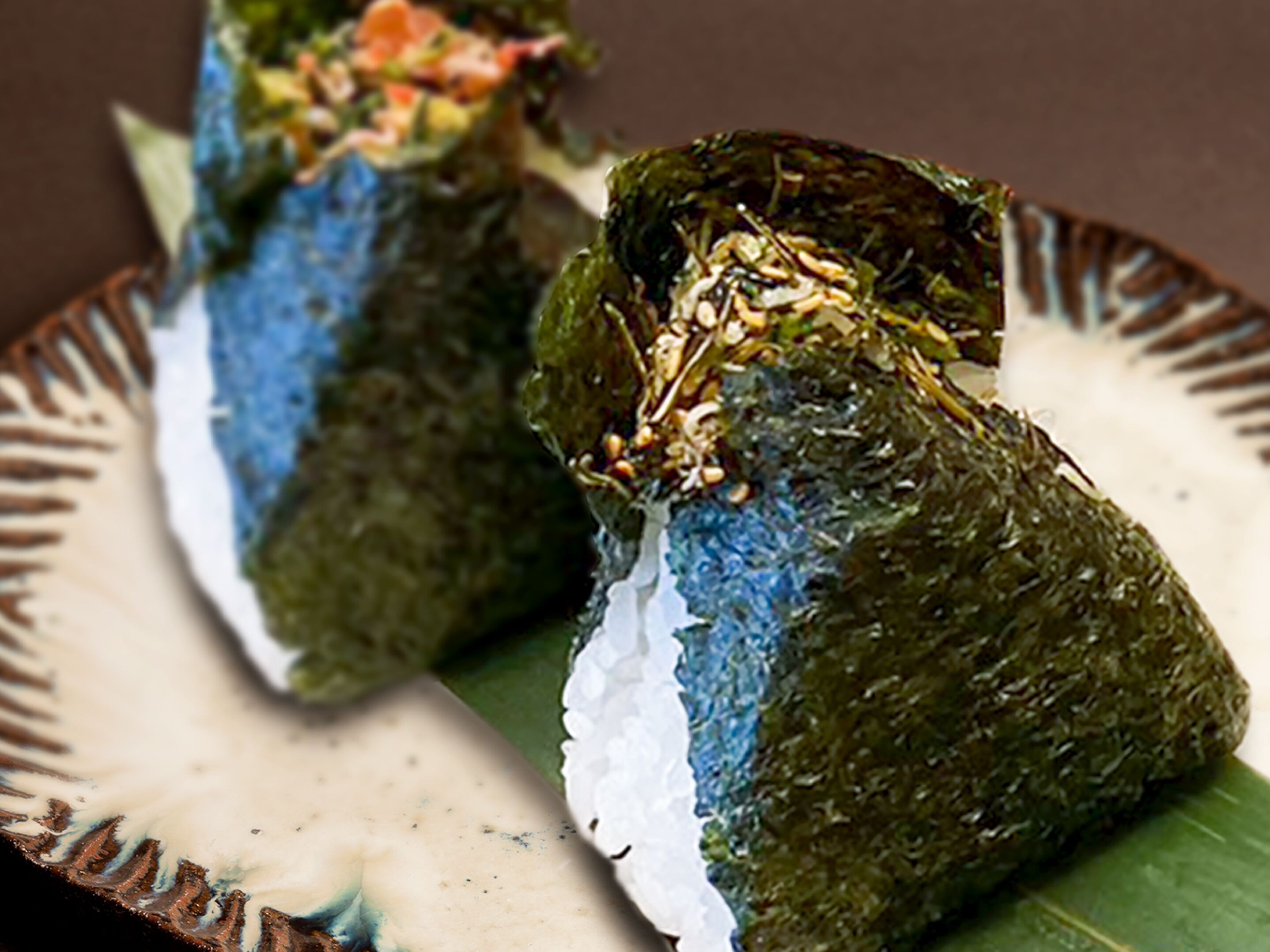 A plate with two sushi rolls, one with a blue topping and the other with a green topping, both with white rice and a garnish on top.