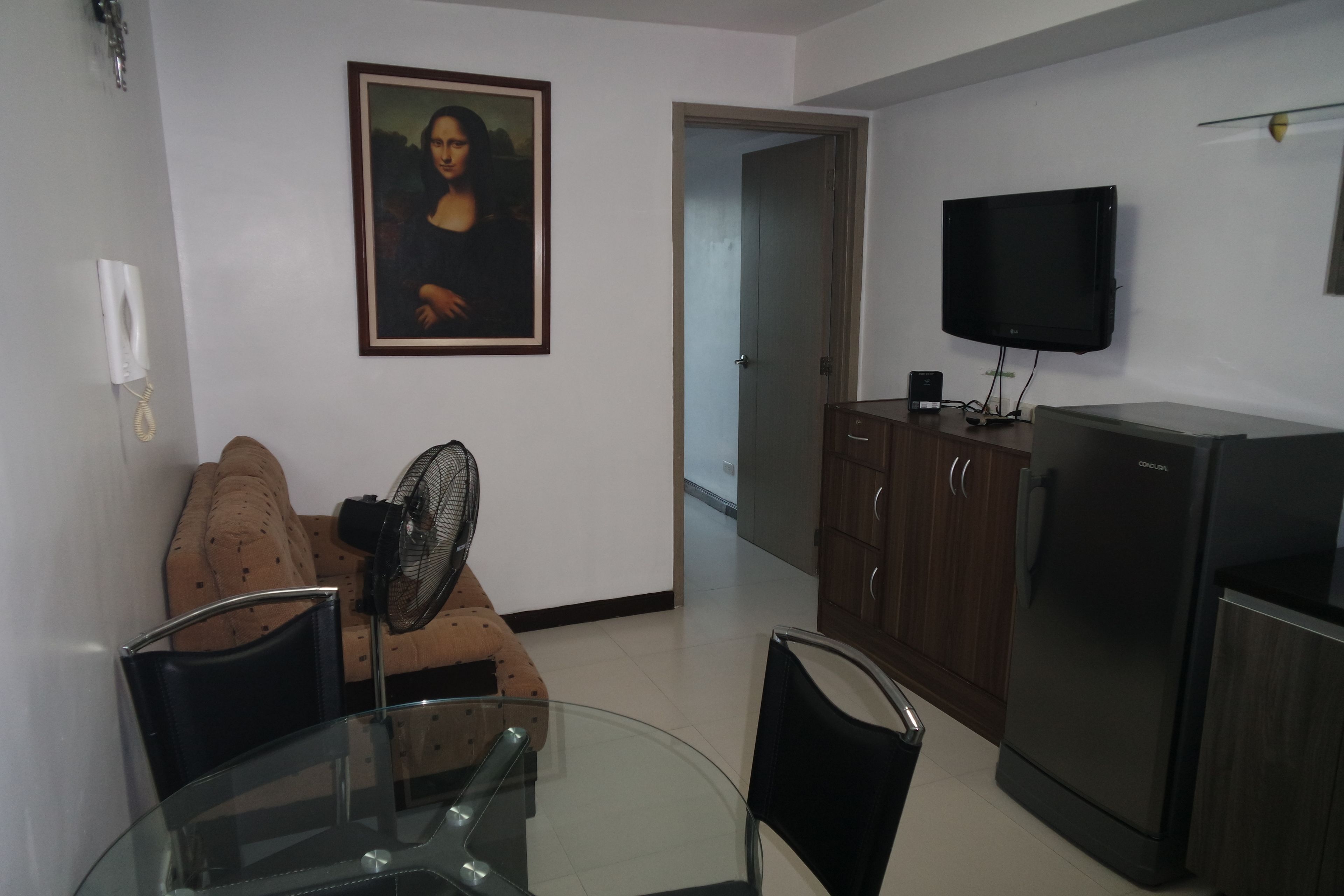 A living room with a glass table, a sofa, a fan, a TV, and a refrigerator.