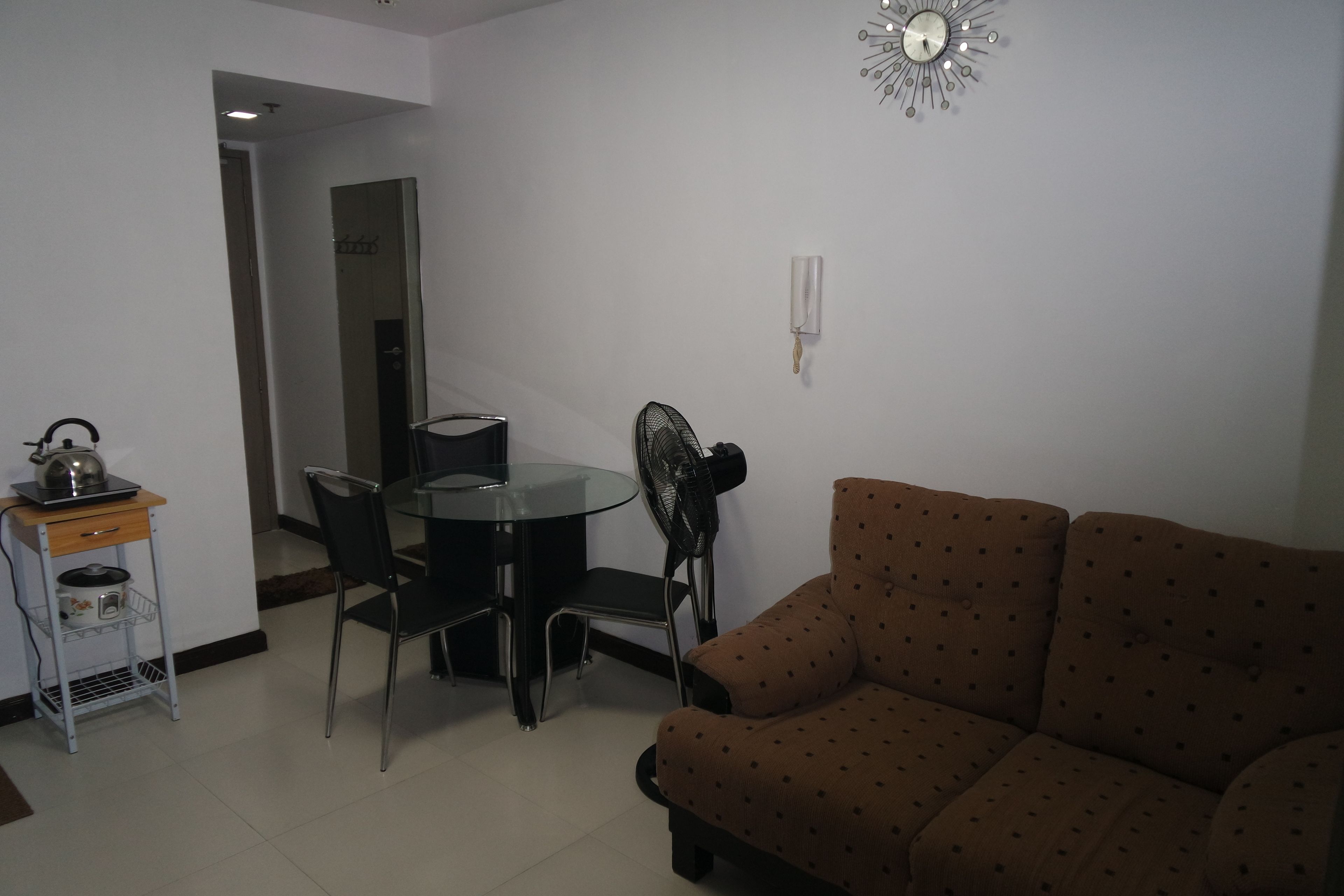 A living room with a brown sofa, a glass table, and chairs. There is a small kitchen area with a kettle and a fan.