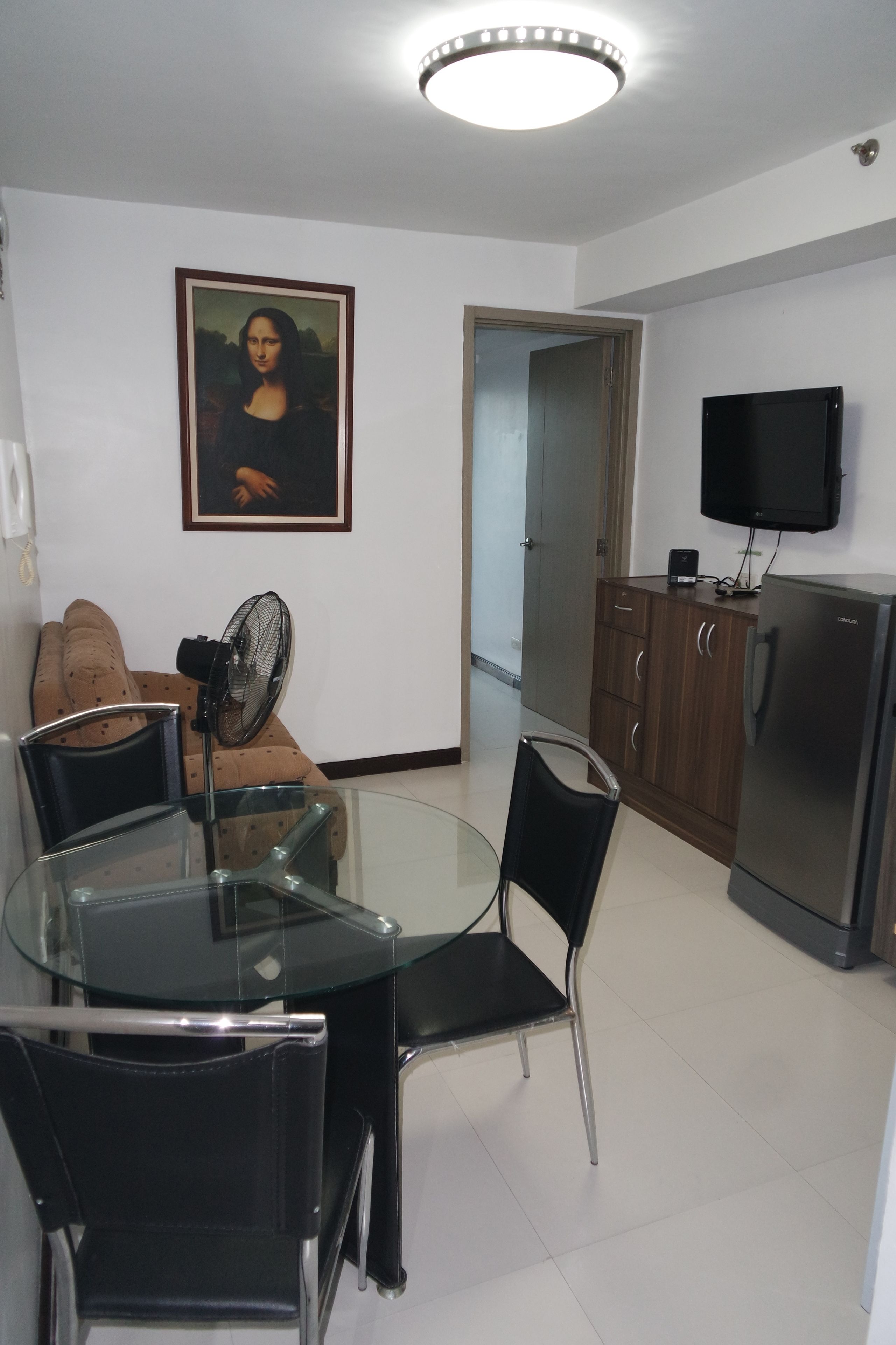 A dining area with a glass table, black chairs, a fan, a television, and a refrigerator.