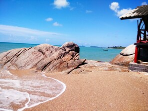 Ein Strand mit großen Felsen, einem kleinen Gebäude mit rotem Dach und ruhiger See.
