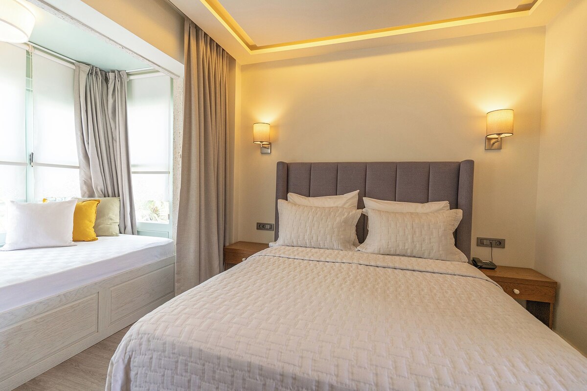 A hotel room with a bed, bedside tables, a window with curtains, and wall-mounted lighting.