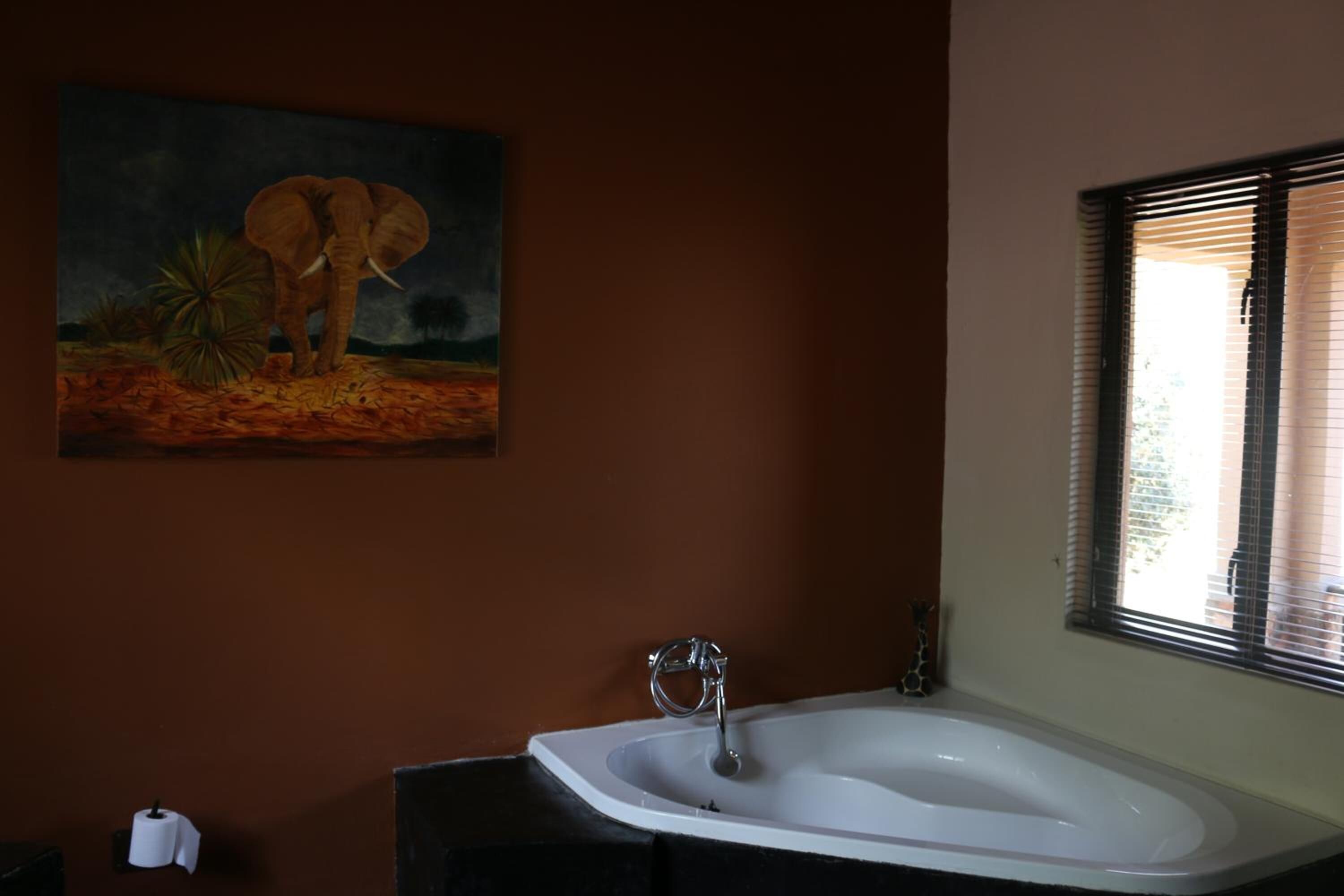 A bathroom with a white bathtub, a painting of an elephant, and a window with blinds.