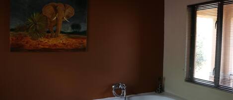 A bathroom with a white bathtub, a painting of an elephant, and a window with blinds.