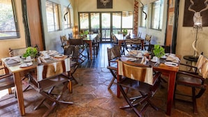 Un comedor con varias mesas y sillas, piso de piedra y ventanas amplias.