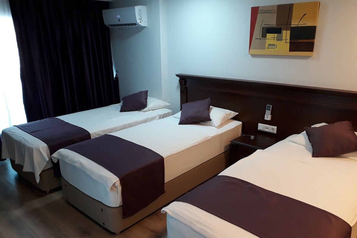 Hotellihuoneessa on kolme erillistä sänkyä, joissa kaikissa on valkoinen patja ja ruskea peitto. Huoneessa on seinään asennettu ilmastointilaite, yöpöytä lampun kanssa ja seinällä on taulu.