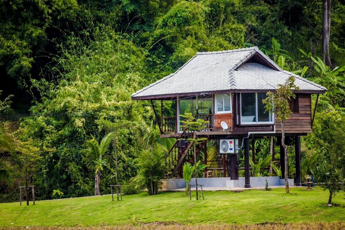 A wooden house with a tiled roof, elevated on stilts, surrounded by lush greenery.