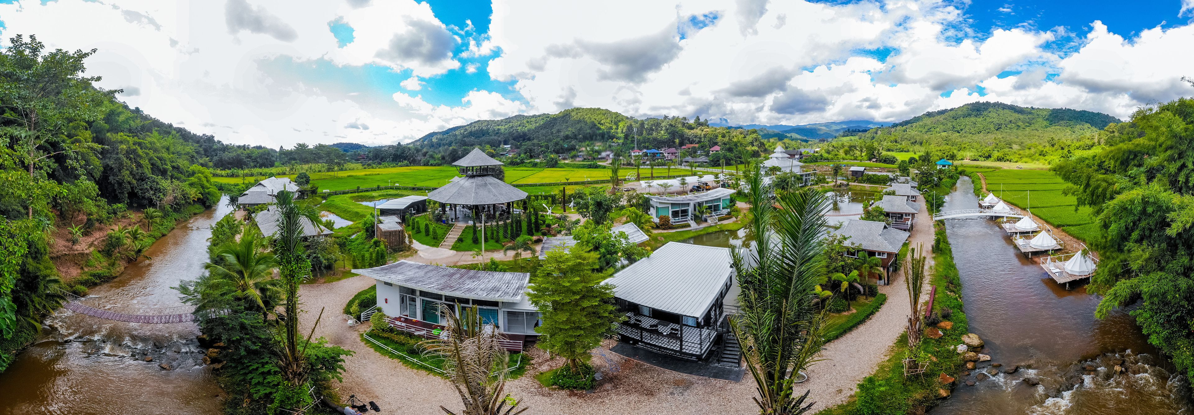 A riverside resort with multiple buildings, including a central pavilion, guest houses, and a bridge over the river.